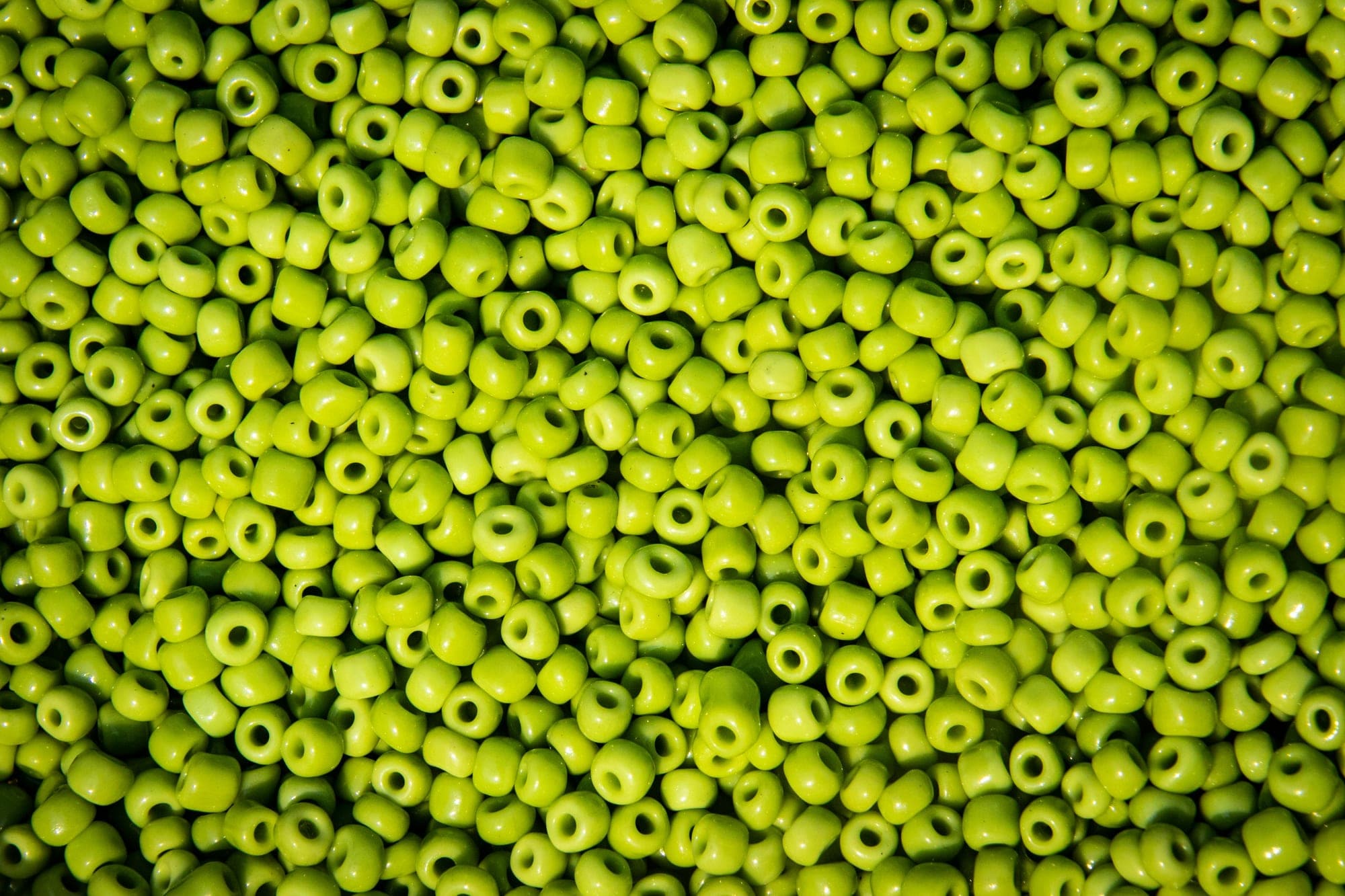 A dense field of glass seed beads in yellow-green, filling the frame. Each bead is a few millimetres across, and the light catches a different face of every one.